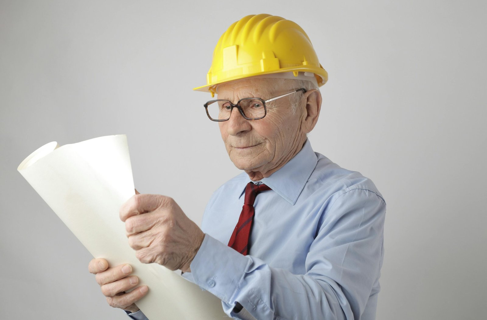 Thoughtful aged male contractor wearing formal shirt and construction helmet examining paper draft
